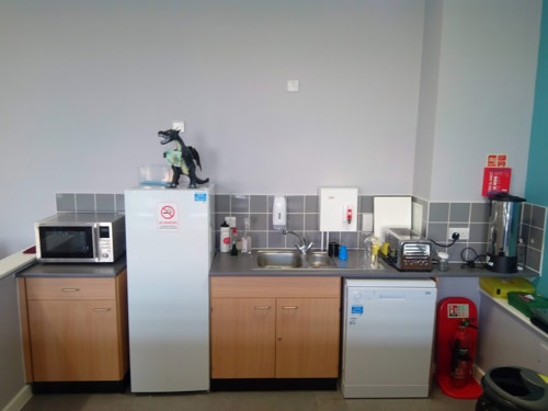 This is an image of the kitchenette in the Cafe.  There is a microwave, dishwasher, toaster, hot water urn, fridge and model dragon holding a sign that says "welcome to the wing"!