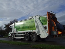 The Council's first electric bin lorry. It has a green paint job that contains trees, buildings with solar panels, the sun, wind turbines and other similar icons