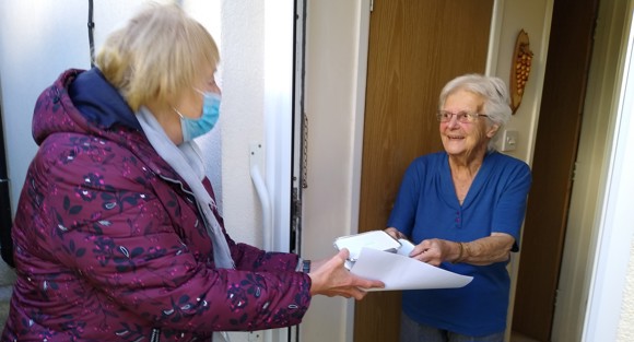 Elderly lady in blue at door receiving a meal from volunteer in mask