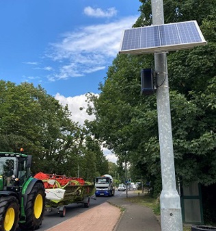 A Zephyr air quality monitor and solar panel attached to a lamppost next to a road with traffic including a tractor