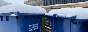 Snow sitting on top of some blue wheelie bins.