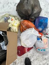 Household waste dumped on snow-covered ground. The waste includes a full black dustbin bag, a cardboard box full of rubbish and several smaller plastic shopping bags that are full of general household waste.