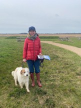 Cllr Bridget Smith and her dog, named Frankie, walking on grass at the Ouse Fen Nature Reserve. There is a light brown path and picnic tables in the background, with a cloudy sky overhead.