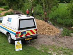 A white Council pick-up truck parked alongside the River Mel. There are two people to the side of the van, looking at conservation work that has taken place on the riverbed.