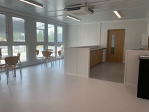  The interior of the Northstowe temporary community centre. This area shows tables and chairs on a white floor and a space for preparing food.
