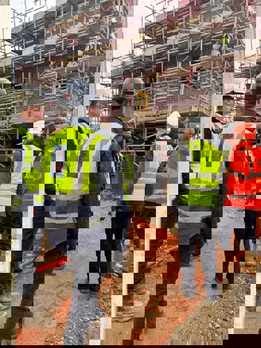 A group of people in high‑visibility clothing and protective helmets stand on a construction site looking towards scaffolding surrounding a building under construction. Some construction workers are positioned on the scaffolding above.