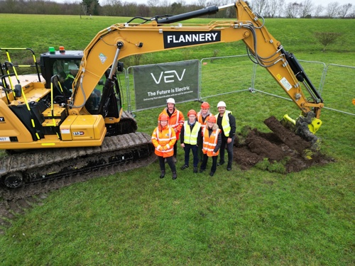 A group of six people wearing high-visibility jackets and hard hats stand on a grassy field next to a large yellow excavator with the name “FLANNERY” on its arm. The excavator’s bucket is digging into the ground, lifting soil. Behind the group is a temporary metal fence with a banner that reads “VEV – The Future is Electric – Delivering Net Zero for Fleets.” The background shows an open green landscape with trees in the distance under a cloudy sky.