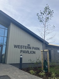 The outside of the Northstowe Western Park Pavilion. The building has a pitched roof and there are new shrubs and trees in the foreground.