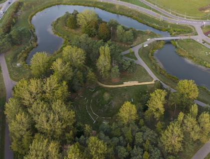 Aerial view of a park in Waterbeach new town featuring two ponds surrounded by trees and greenery. A bridge connects the ponds, and winding pathways allow visitors to explore the area. Roads border the park on two sides, providing vehicle access.